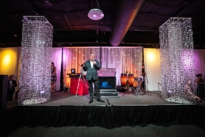 Man on Stage with Flanking Silver Beaded Columns and a LED Star Drop / Star Curtain Background with an uplit White String Drapery From Turn of Events Las Vegas Rental Drapery