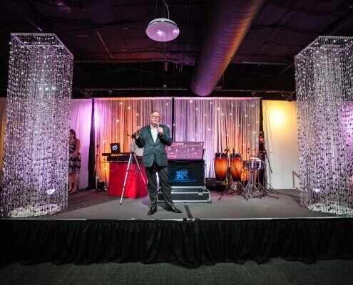 Man on Stage with Flanking Silver Beaded Columns and a LED Star Drop / Star Curtain Background with an uplit White String Drapery From Turn of Events Las Vegas Rental Drapery